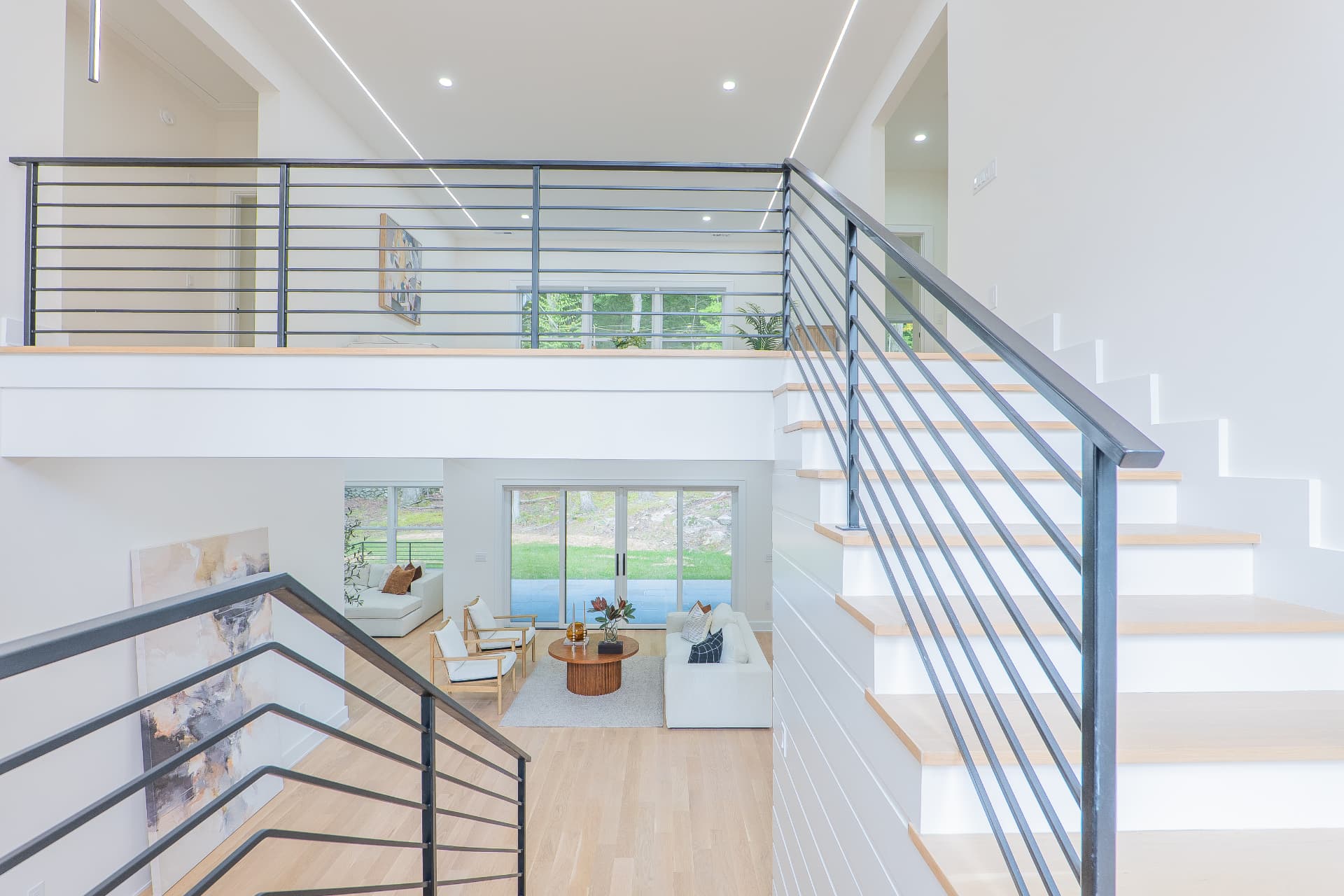 Staircase with blackened steel railing and built-in track lighting above a double-height living room